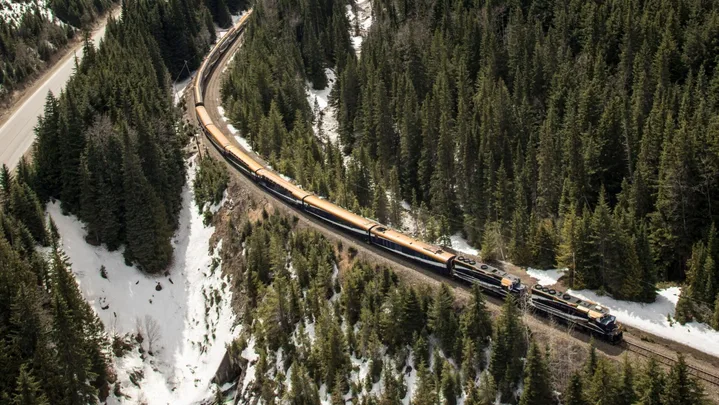 Aerial view of a train traveling through a snowy mountain landscape with pine forests and a towering peak in the background.