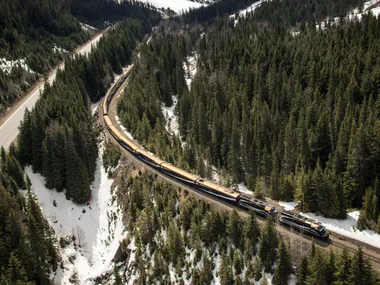 Aerial view of a train traveling through a snowy mountain landscape with pine forests and a towering peak in the background.