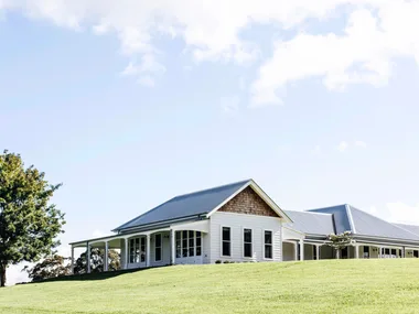 Single-story white house with a gray metal roof, surrounded by green lawn and a tree, under a blue sky.