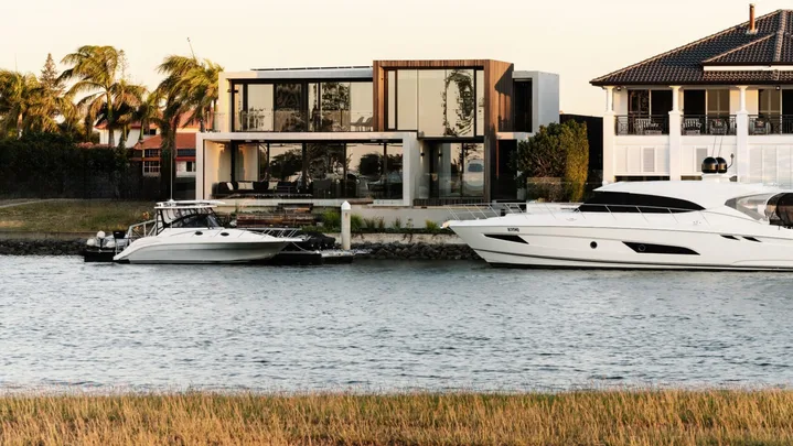 Modern canalside house with large glass windows, palm trees, and two yachts moored by the dock at sunset.