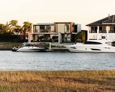 Modern canalside house with large glass windows, palm trees, and two yachts moored by the dock at sunset.