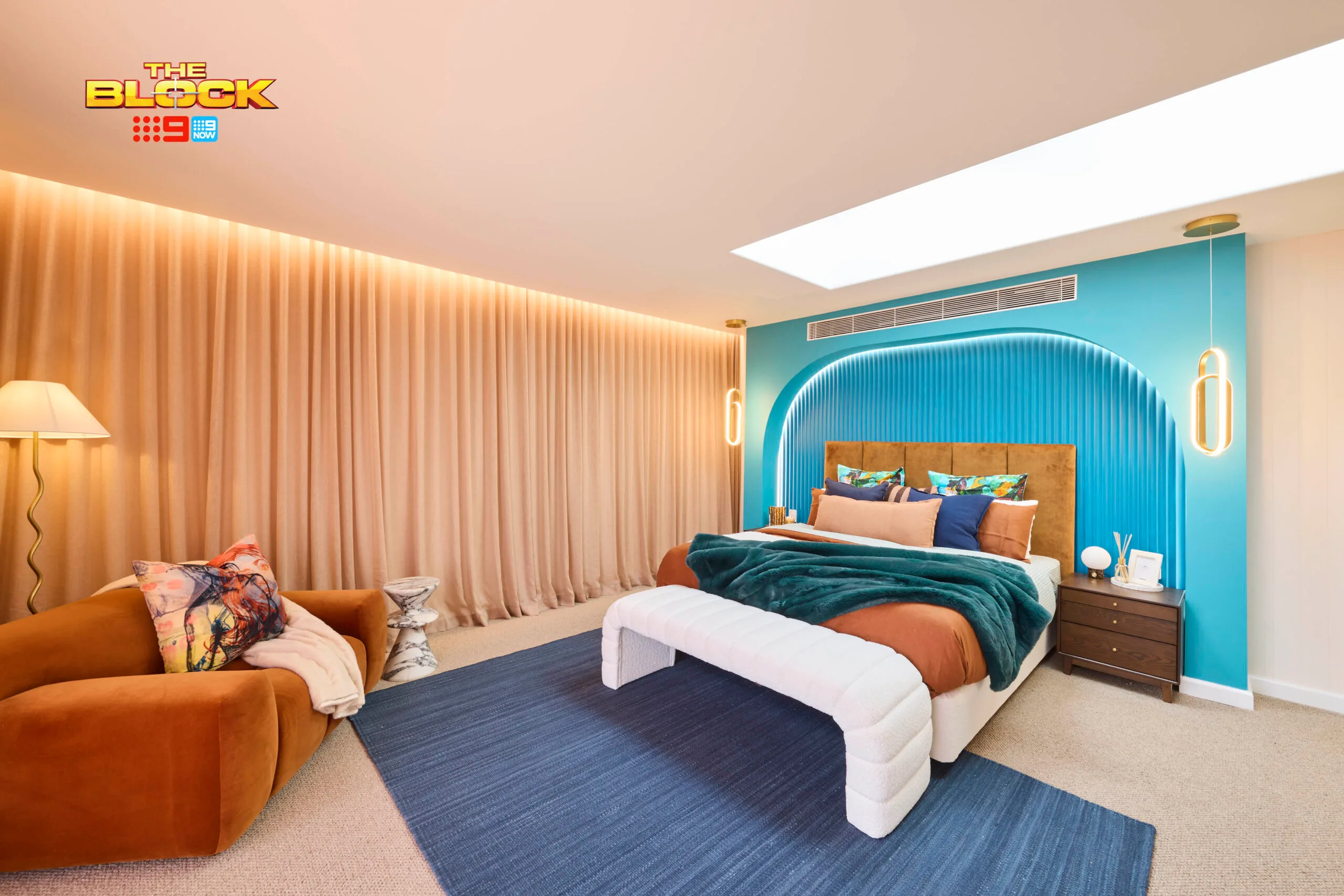A modern bedroom from "The Block" with a teal accent wall, velvet sofa, and ample lighting.