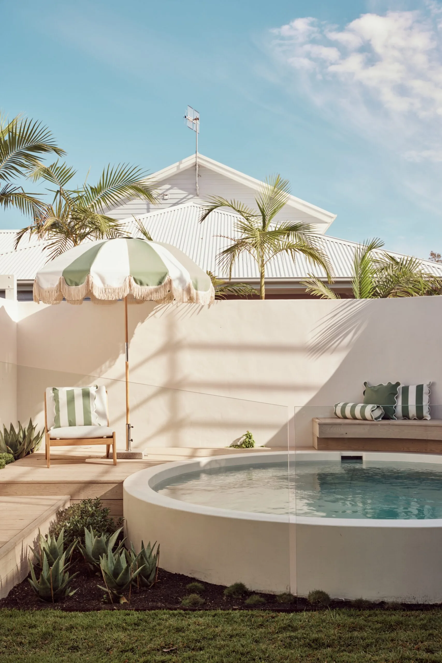 Tropical backyard with small round plunge pool, green and white striped umbrella, chair, palm trees, and a clear sky.
