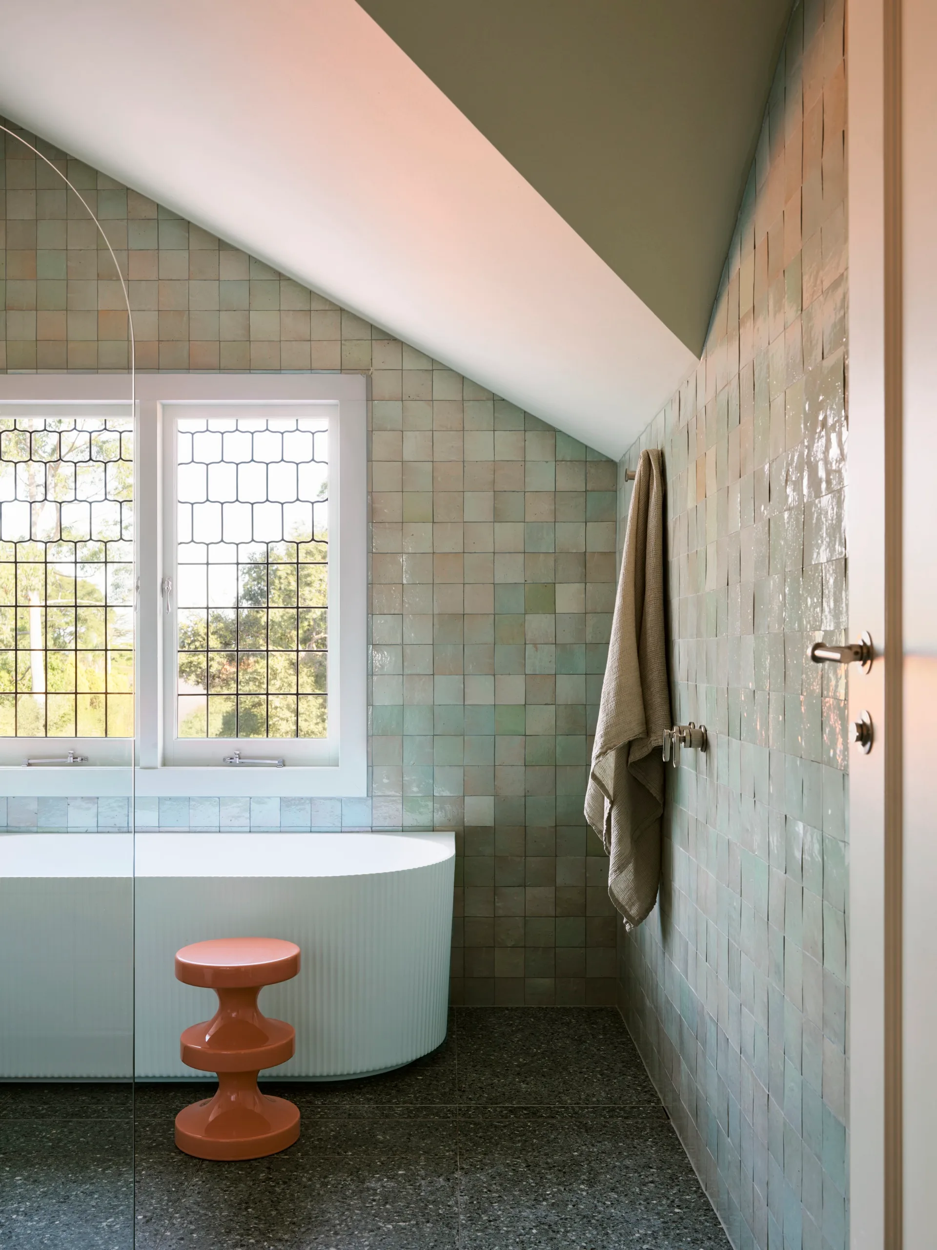 Bright bathroom with green tiled walls, sloped ceiling, white bathtub, orange stool, and window with grid pattern glass.