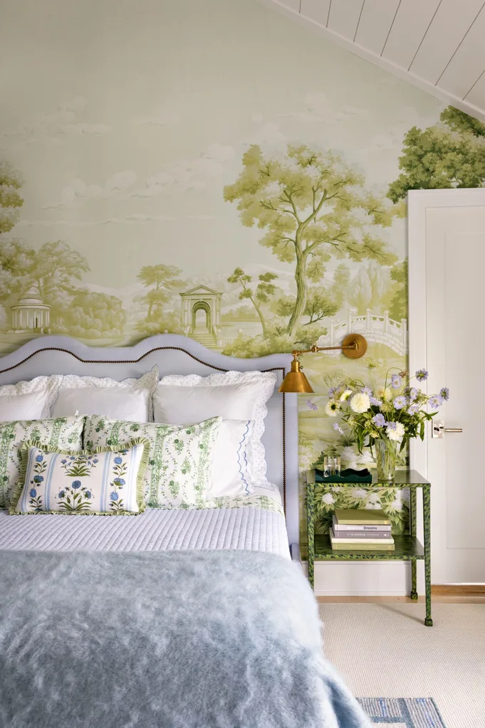 Bedroom with pastoral mural wallpaper, bed with floral pillows, blue throw, and a side table with flowers and books.