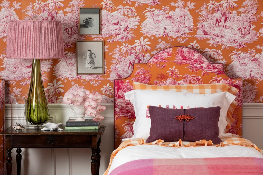 Cozy bedroom with vibrant orange and pink wallpaper. The bedhead is upholstered in the same fabric. A green glass lamp, and floral accents on a wooden side table.