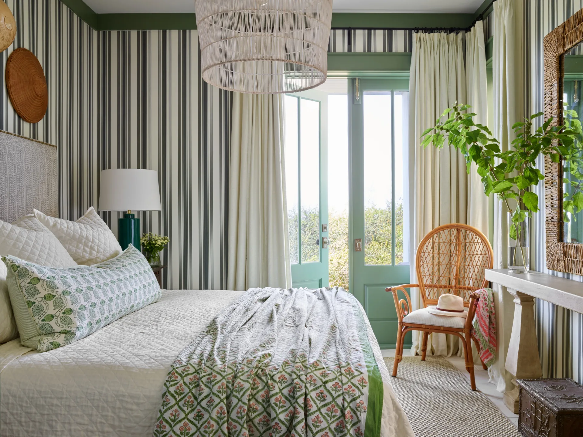 Cozy bedroom with striped walls, green accents, wicker chair, and a bed with patterned bedding near green doors with window panes leading to the outside area.