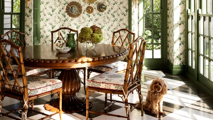 Elegant dining room with green lattice ceiling, floral wallpaper, round table, bamboo chairs, and a small dog on checkered floor