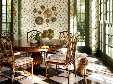 Elegant dining room with green lattice ceiling, floral wallpaper, round table, bamboo chairs, and a small dog on checkered floor