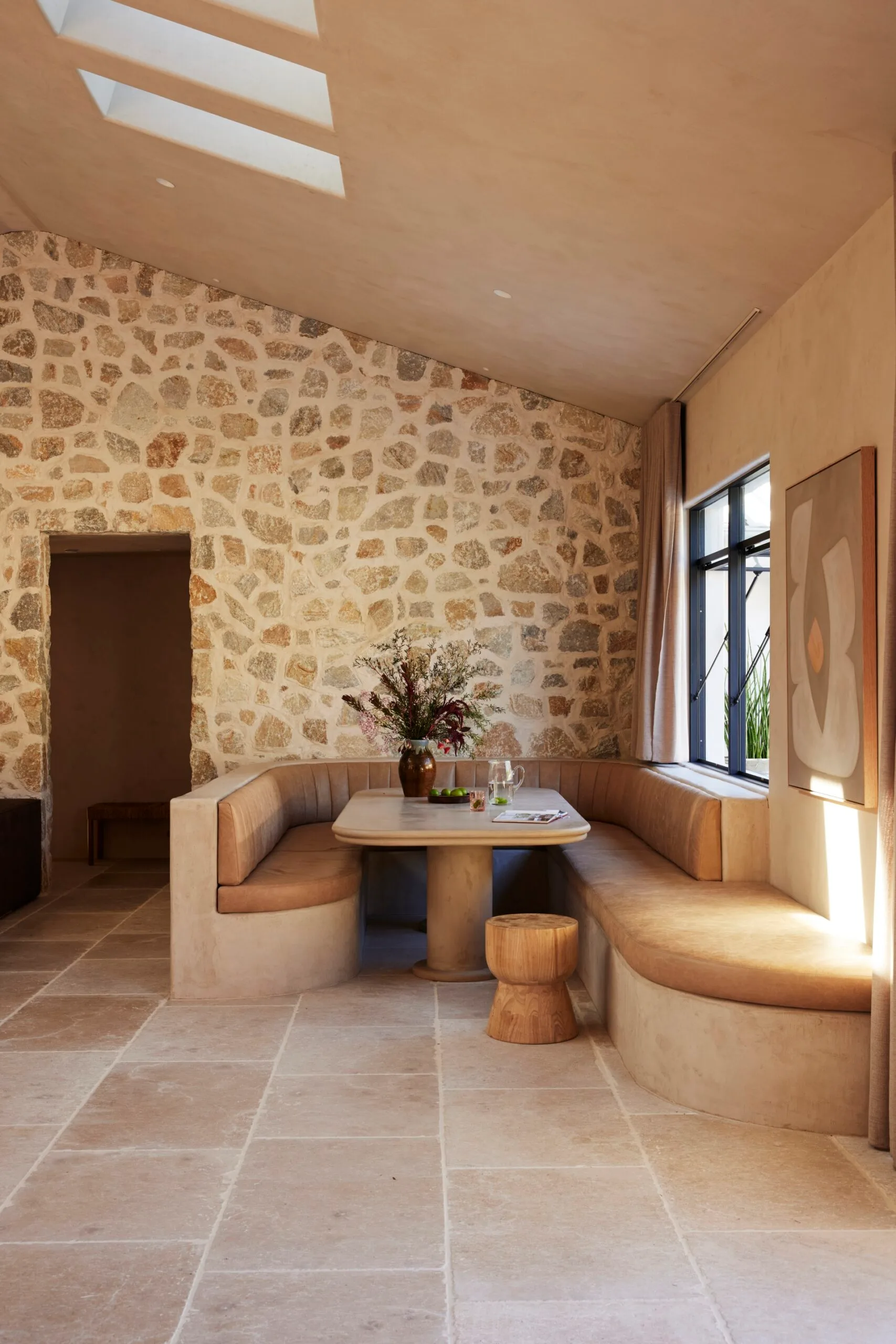 Cozy dining nook with built-in banquette seating, stone wall, skylights, and a window, featuring a vase of flowers on the table. Dining room ideas.
