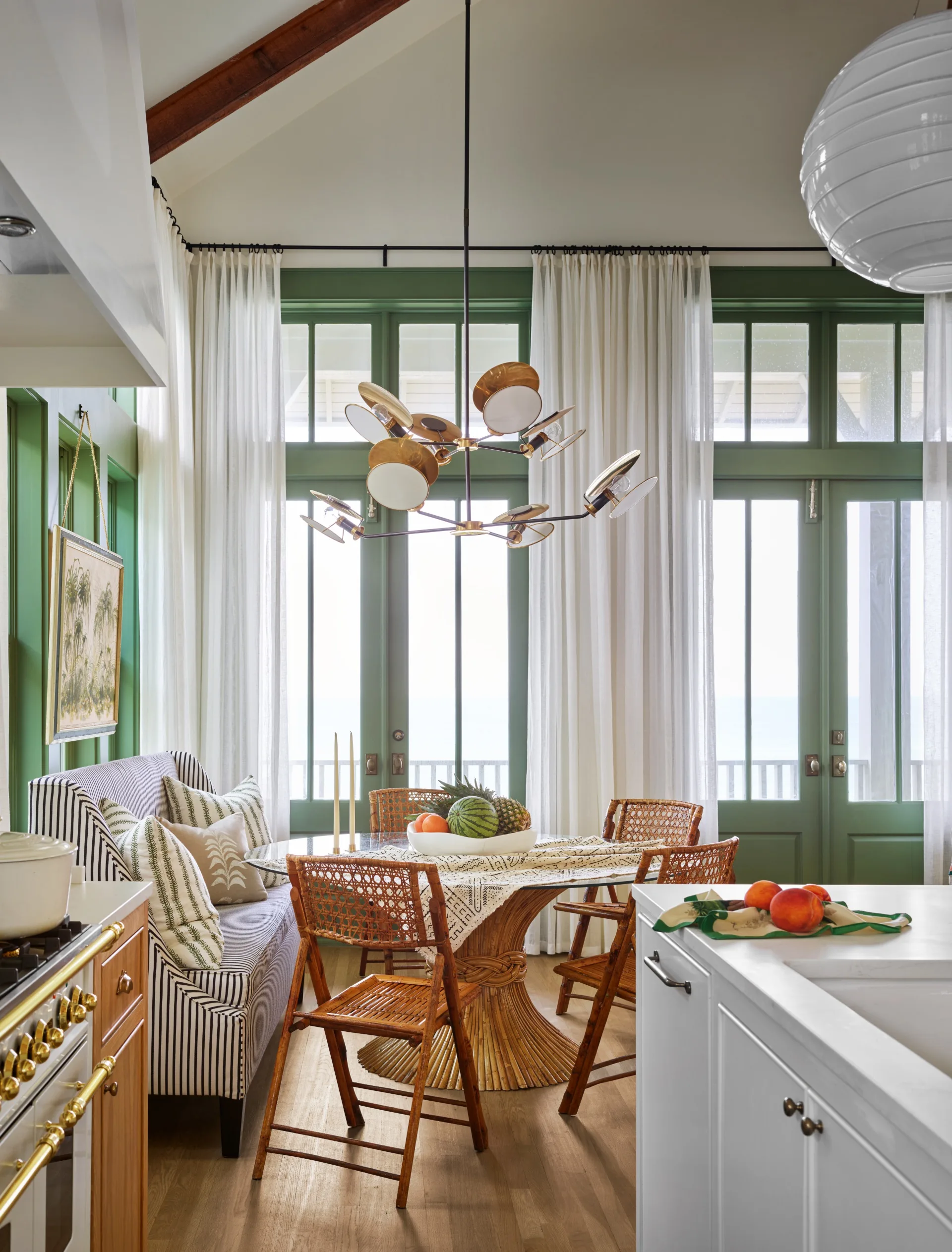 Elegant kitchen with a breakfast nook that has wicker chairs, a round table, striped banquette, and modern chandelier, overlooking a large window. Dining room ideas.