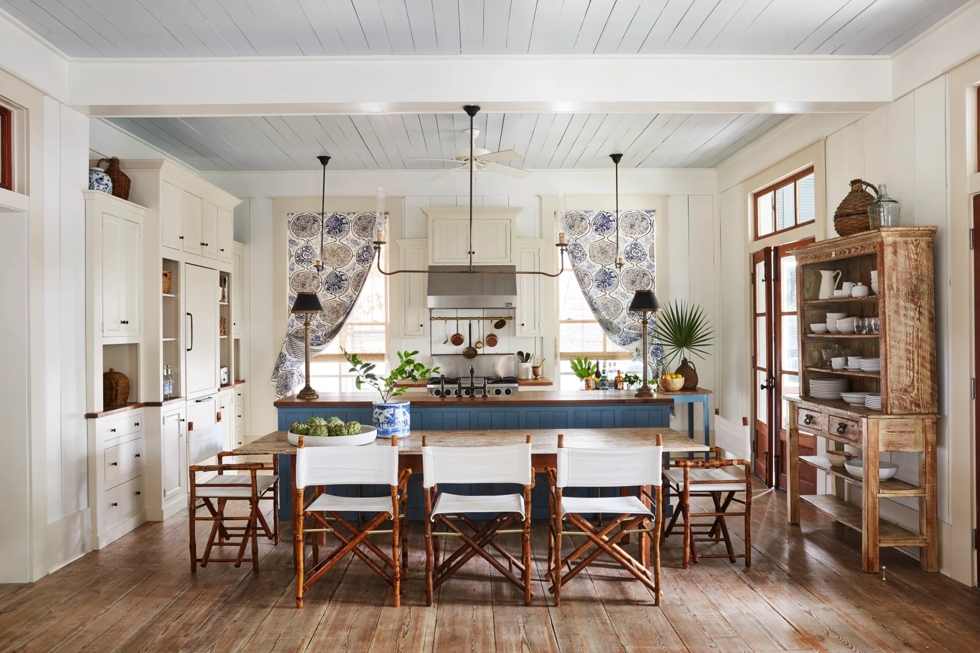 Bright kitchen with wooden floors, white cabinets, patterned curtains, and a large island with blue base and six chairs around a dining table.