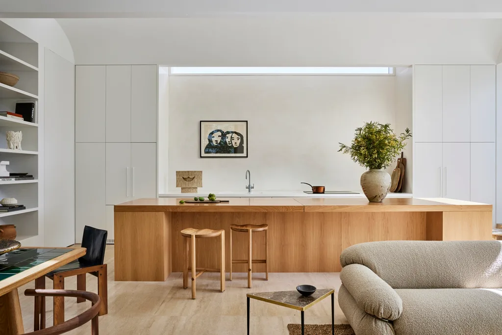 Modern kitchen with wood island, stools, and potted plant. Art on wall, bookshelves and sofa visible