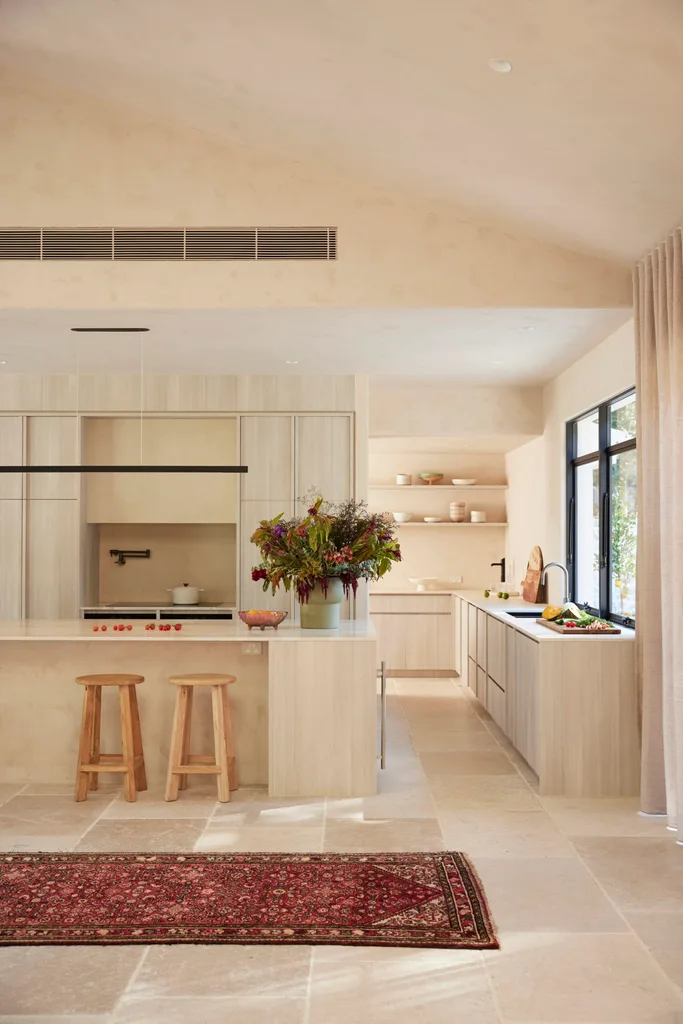 Modern kitchen with light wood cabinets, two stools tucked against the kitchen island, a countertop vase of flowers, and a red patterned rug. A pantry is visible around the side of the kitchen.