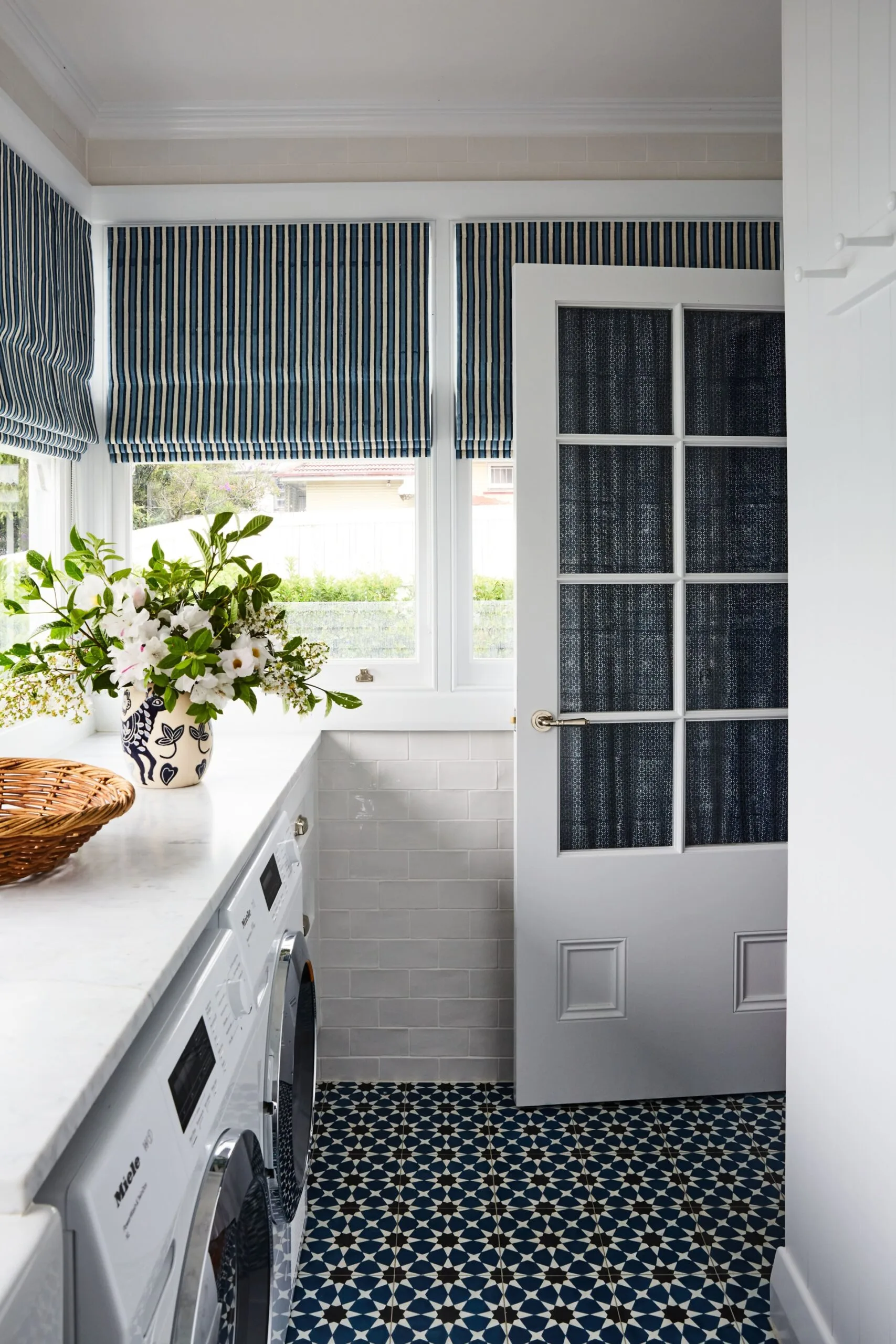 Laundry room with blue-patterned floor tiles, striped Roman blinds, and a vase of flowers on a marble countertop.