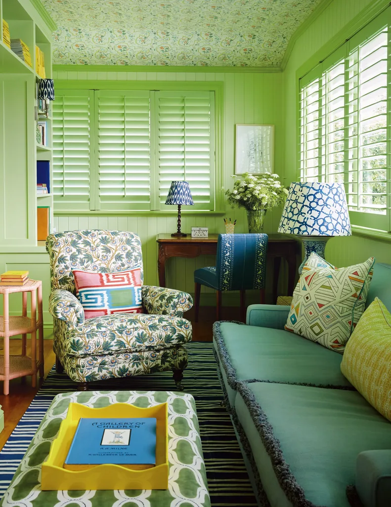 Bright green living room with floral armchair, striped rug, patterned cushions, and decorative lamps. The space is colour drenched, with VJ panelled walls painted green and a green and pink floral wallpaper on the ceilling. Living room ideas.