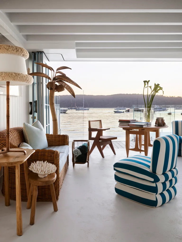 Coastal living room with wicker furniture, striped chair, wooden decor, and large windows showing a bay view with boats.