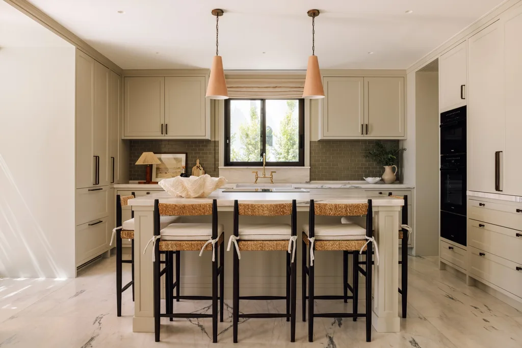 Modern kitchen with beige cabinets, island, four wicker stools, pendant lights, and a window with greenery outside