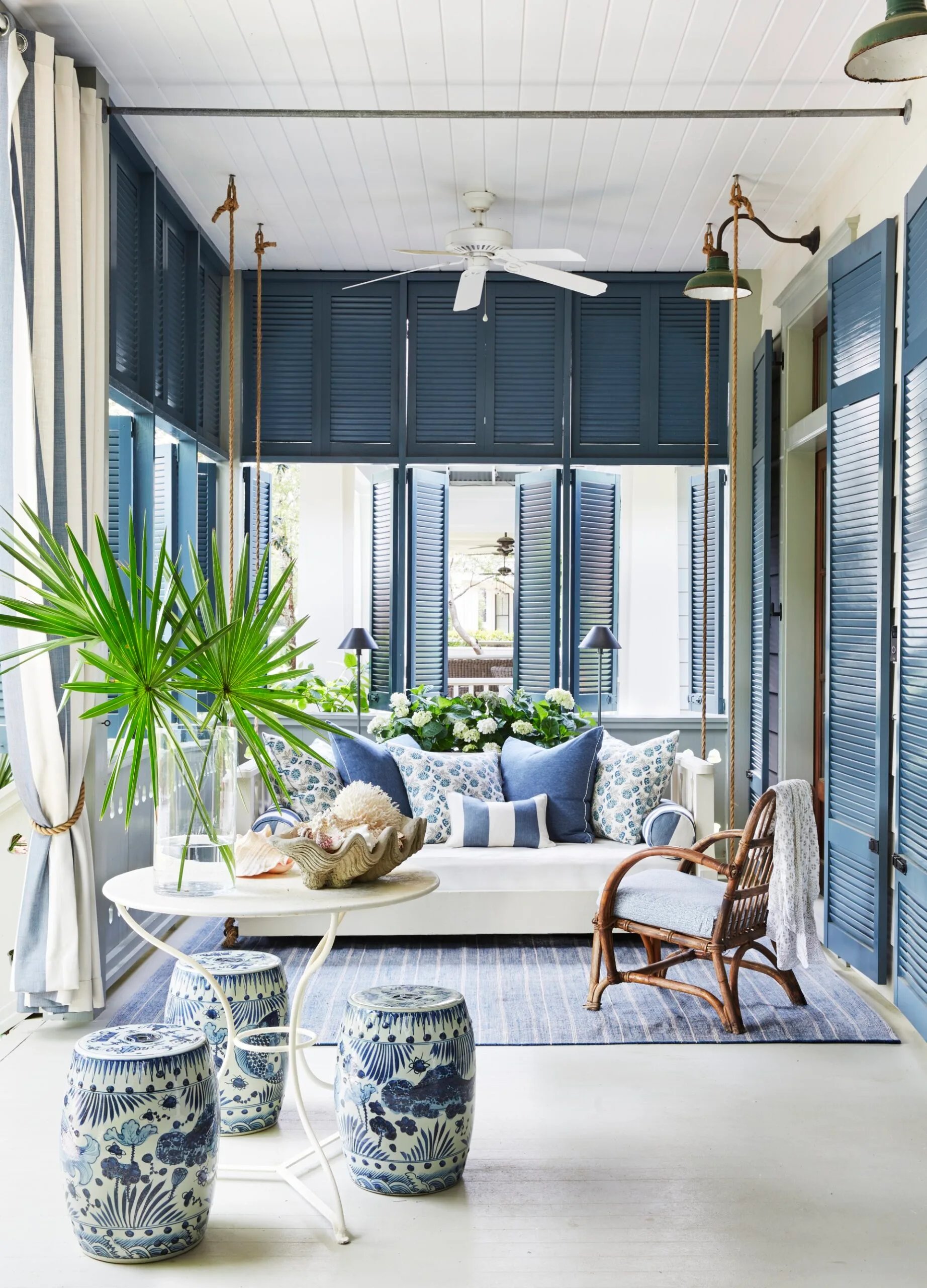 A cozy porch with blue shutters, decorative pillows, a wicker chair, and a table with tropical plants and ceramic stools.  Living room ideas.