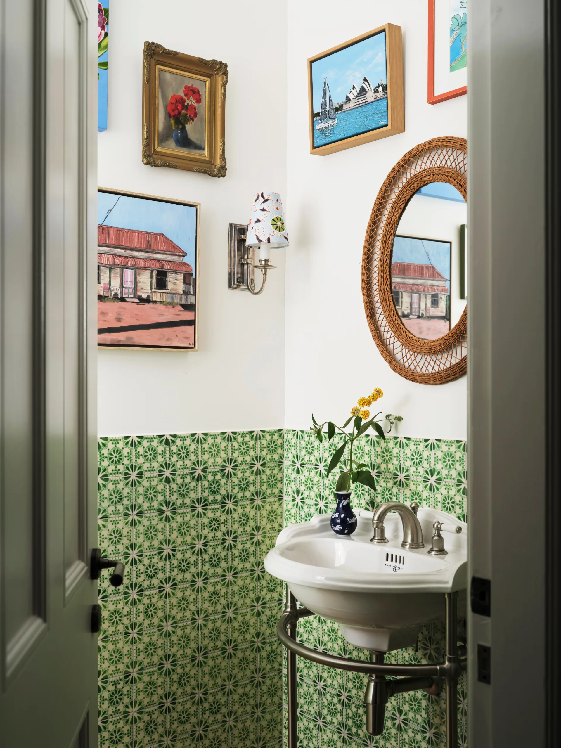 Small powder room with green patterned tiles, framed art, a round mirror, and a white sink with a flower vase