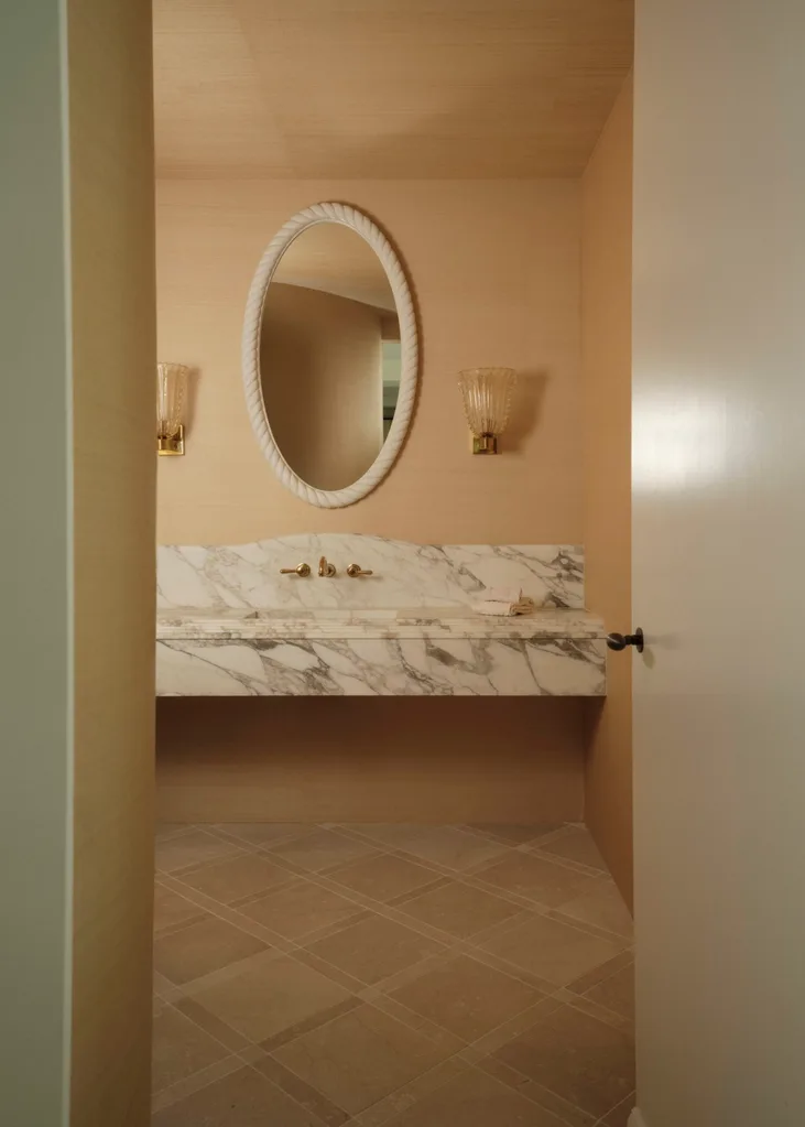 Elegant bathroom with marble sink, oval mirror, and two wall sconces, seen through a slightly open door.
