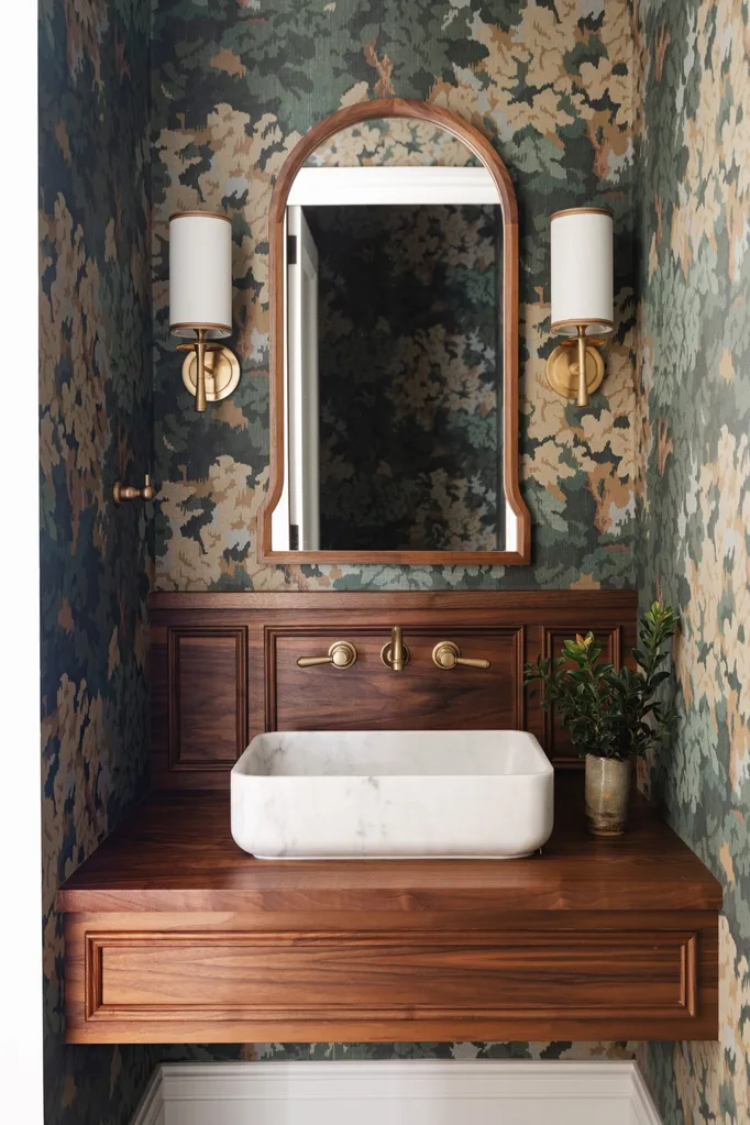 Stylish bathroom with a wooden vanity, marble sink, arched mirror, floral wallpaper, and brass fixtures.