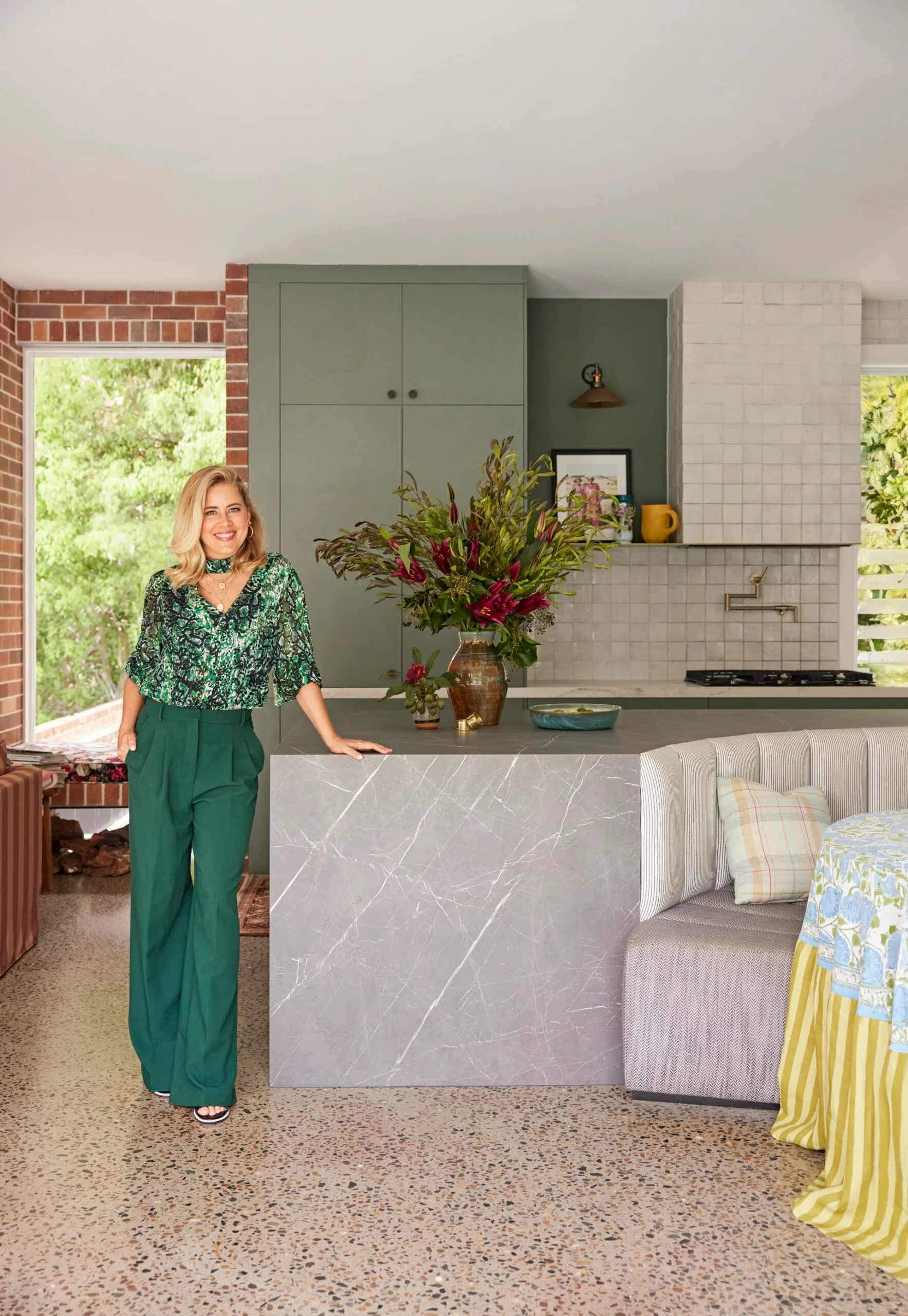 Ex-The Block contestant Carlene Duffy wears green pants and a green shirt and stands in her kitchen, which has square glossy tiles, green cabinetry, grey stone island and a wall of bricks beside it.