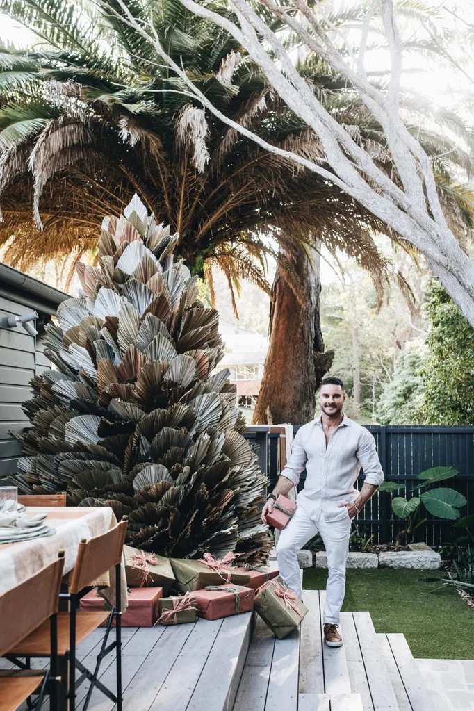 The Block judge Darren Palmer stands on a wooden deck, surrounding by palm trees, with a handmade Christmas tree.