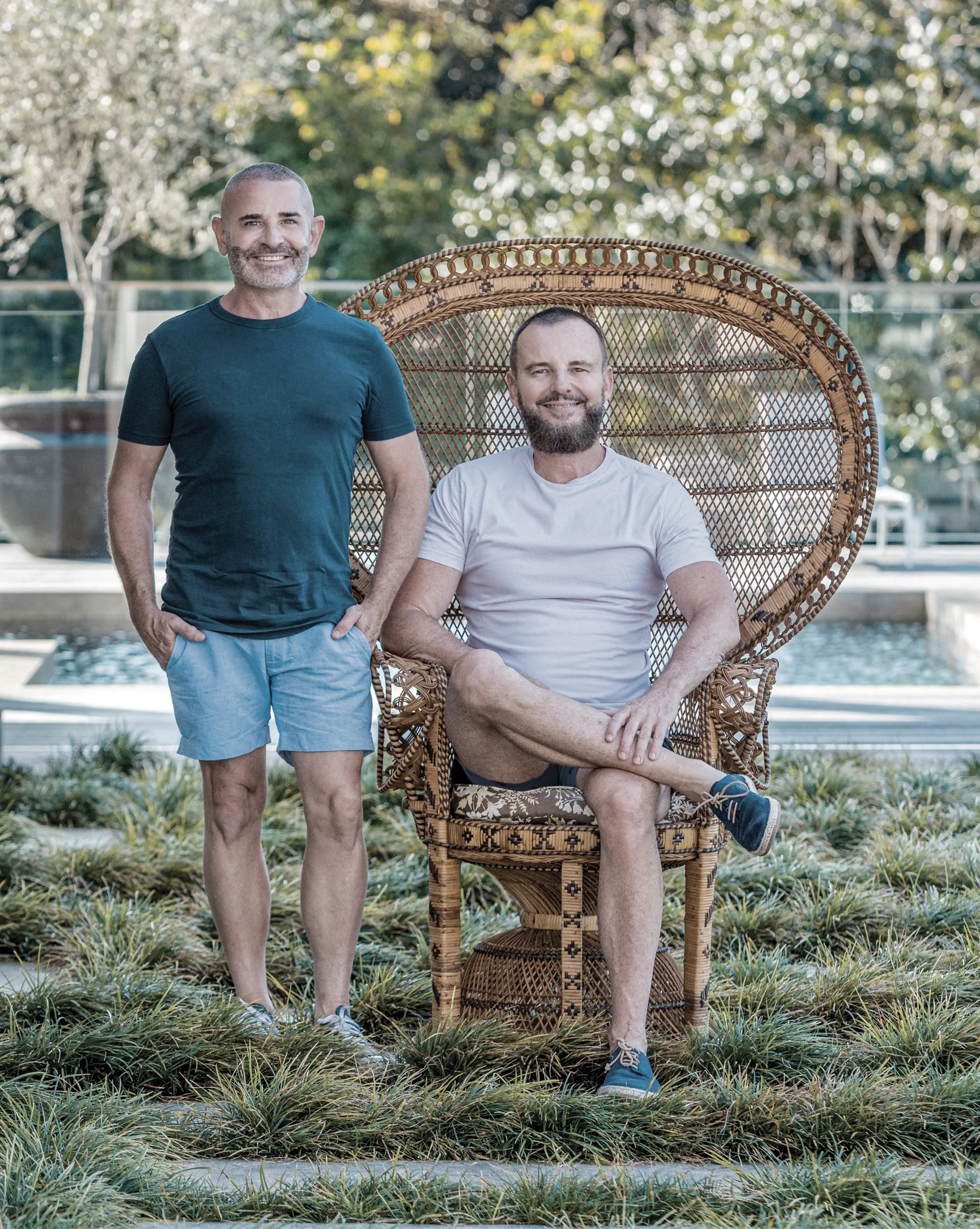 Warren and Gavin from The Designer Boys are outside in casual clothes. One sits on a large rattan charge while the other stands beside.