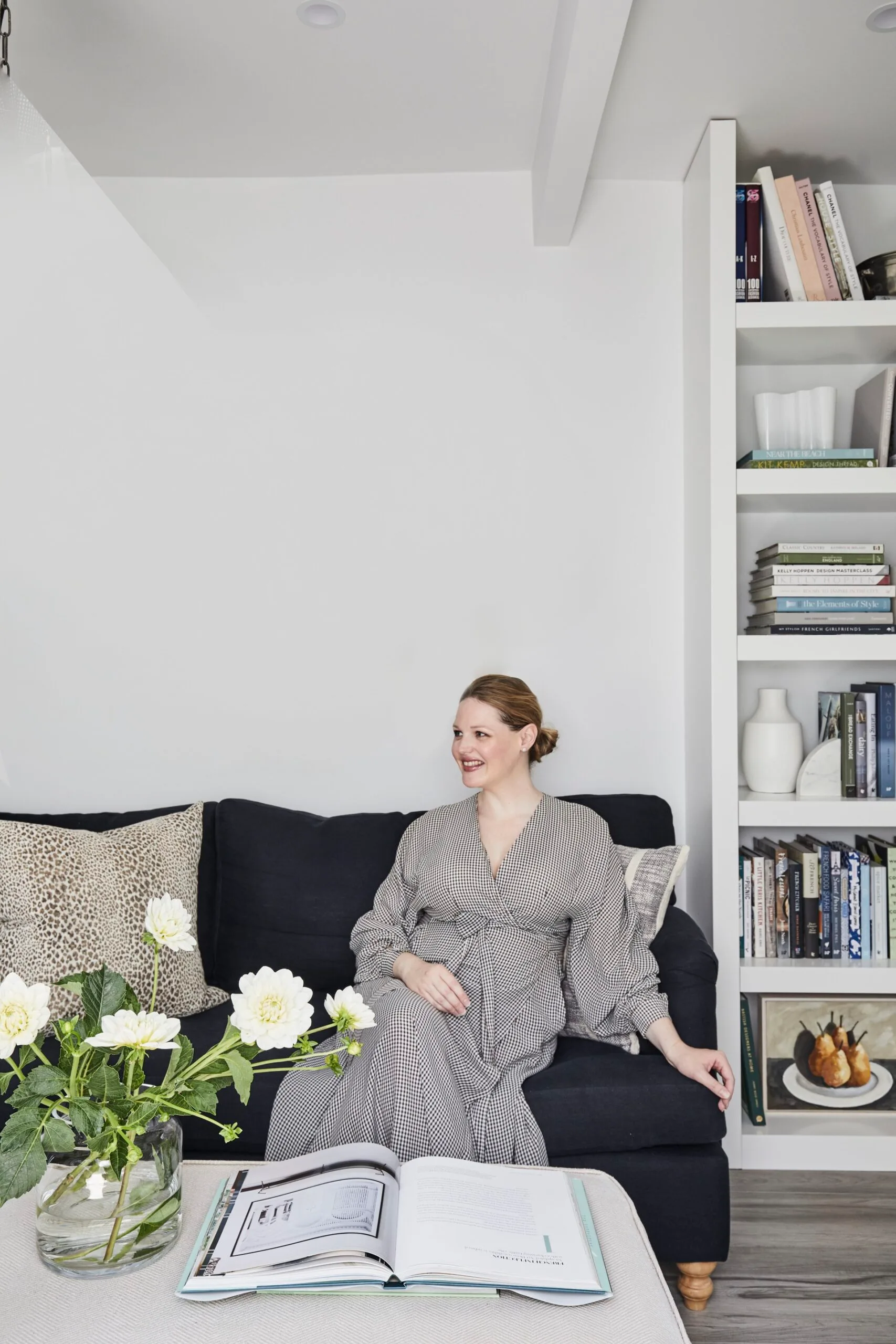 Home Beautiful editor in chief Elle Lovelock sits on a navy sofa. A bookcase is to her left, with a white coffee table in front. A vase of white flowers is positioned on this, next to an open coffee table book.