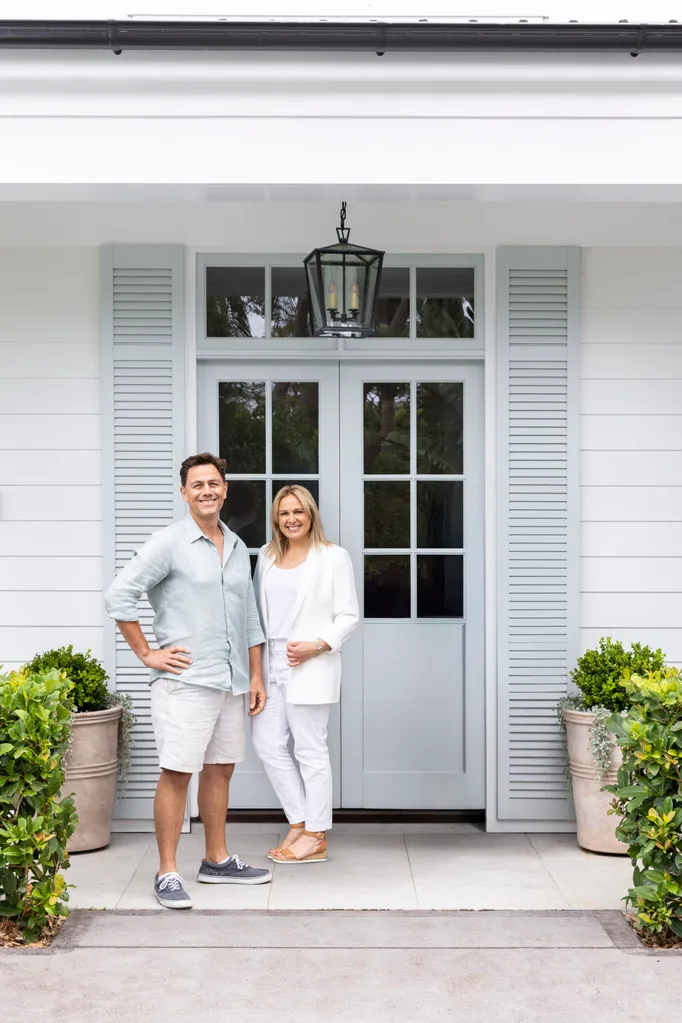 Designers Alex and Janette Stritt stand at white house entrance with light blue shutters and potted plants.