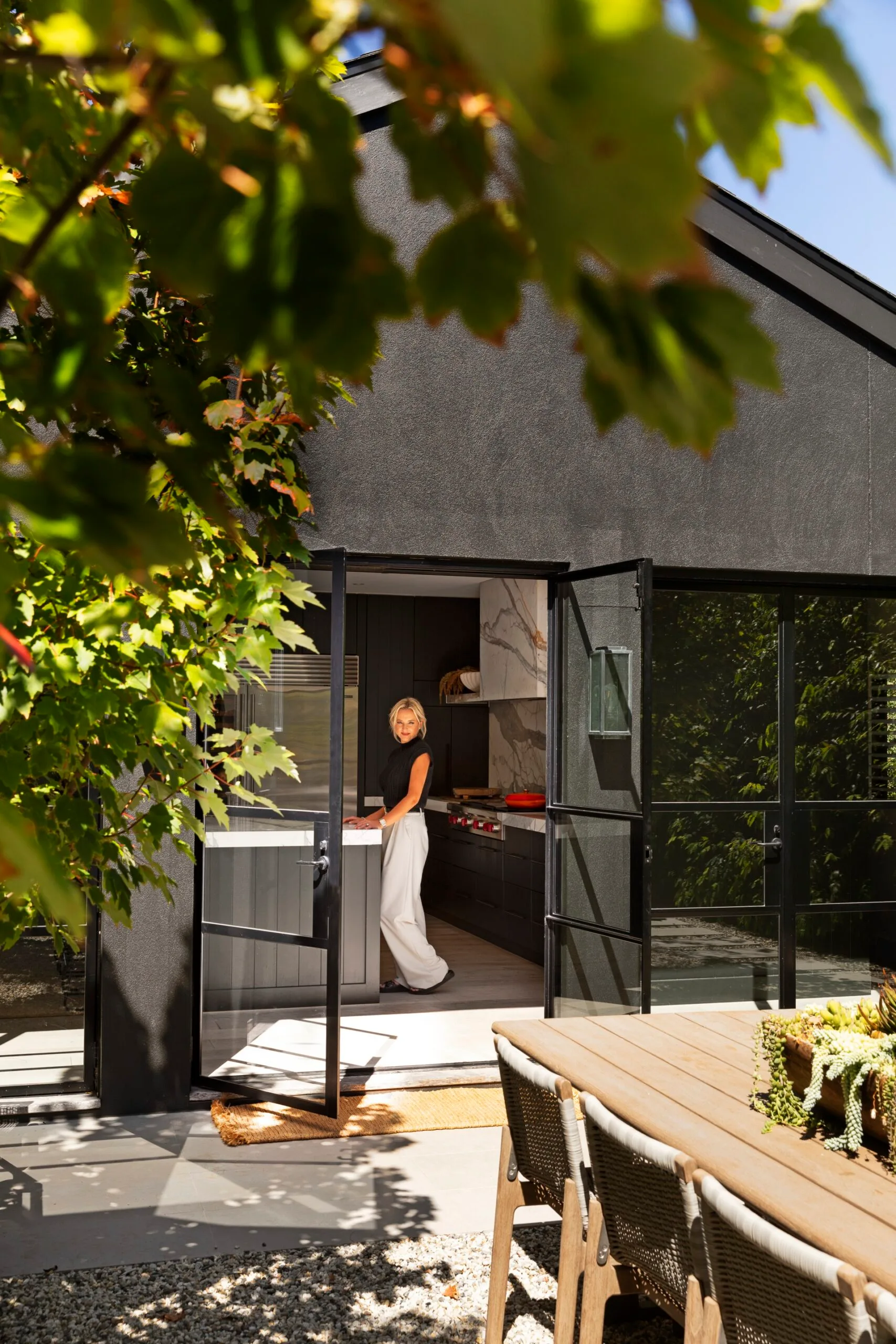 Woman stands in a modern kitchen with glass doors, surrounded by greenery and outdoor dining area. The woman is Australian design influencer Kate Walker.