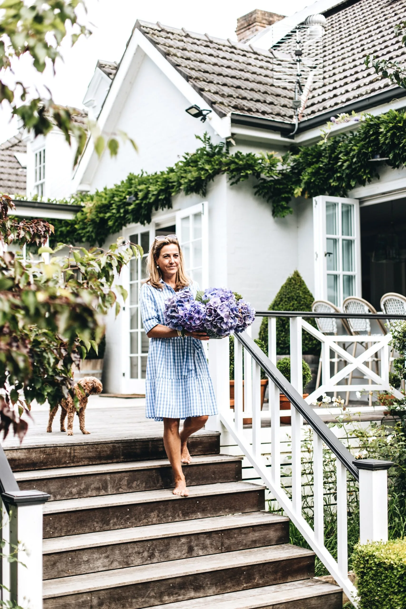 Australian design influencer Melissa Hartwright wears a light blue dress, holds purple flowers while walking down steps of a white house, with a small brown dog nearby.