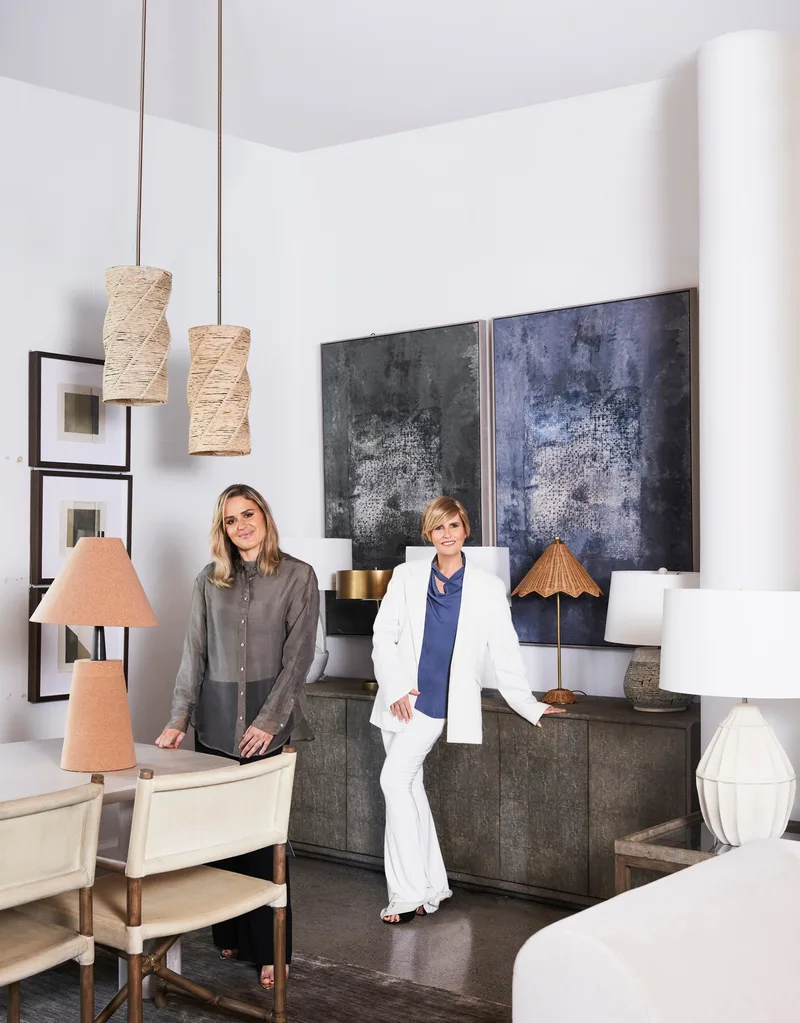 Australian design influencer Melissa Penfold stands in a white suit with a blue shirt, in front of a white wall with two large, dark toned abstract artworks on the wall. Her daughter, Isabella Walker-Smith, stands beside her.
