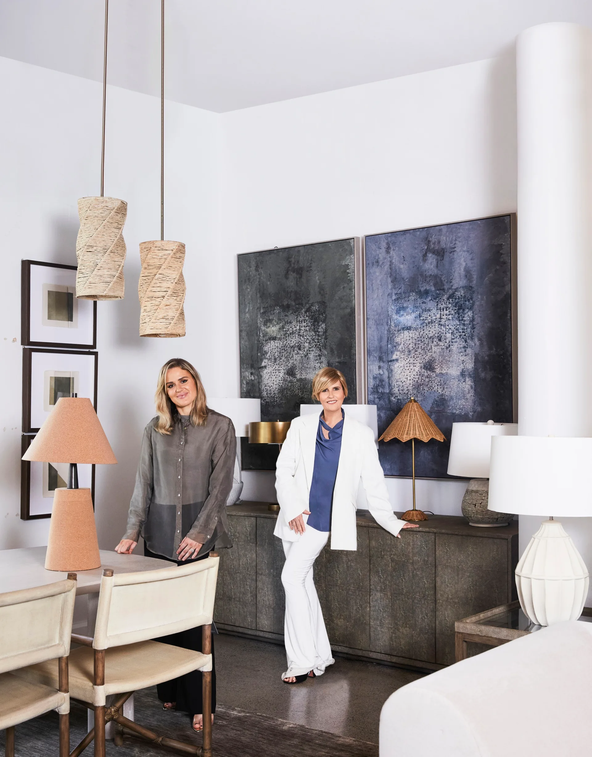 Australian design influencer Melissa Penfold stands in a white suit with a blue shirt, in front of a white wall with two large, dark toned abstract artworks on the wall. Her daughter, Isabella Walker-Smith, stands beside her.