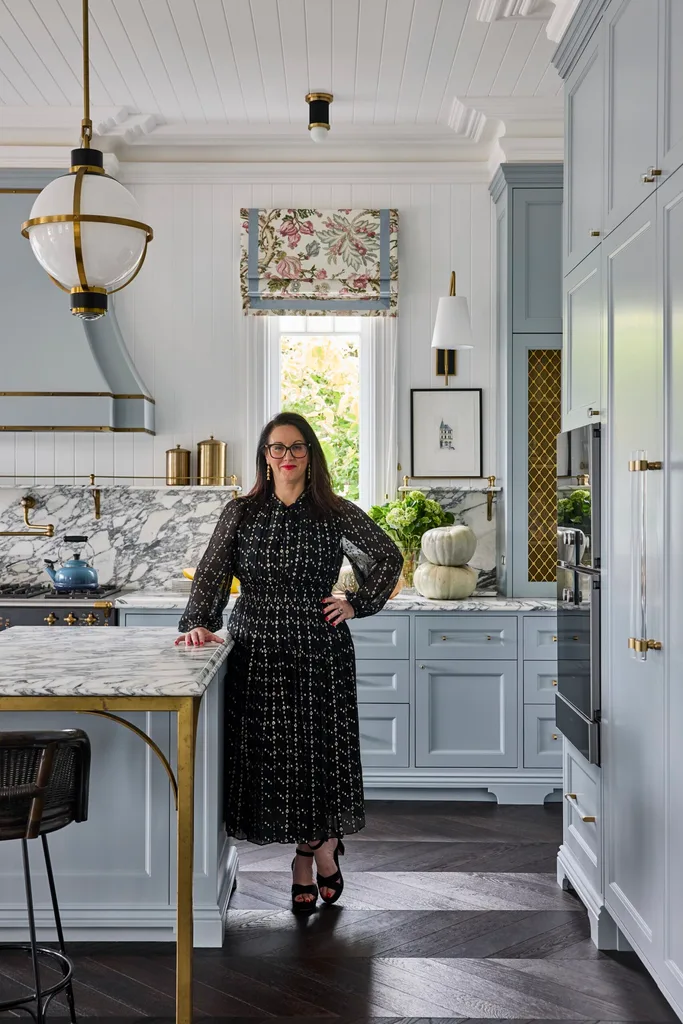 Australian design influencer Lauren Mahoney of Studio Trio stands in her kitchen, which has blue cabinetry, dramatically veined marble benchtops and splashbacks, brass accents and VJ panelled walls and ceiling.