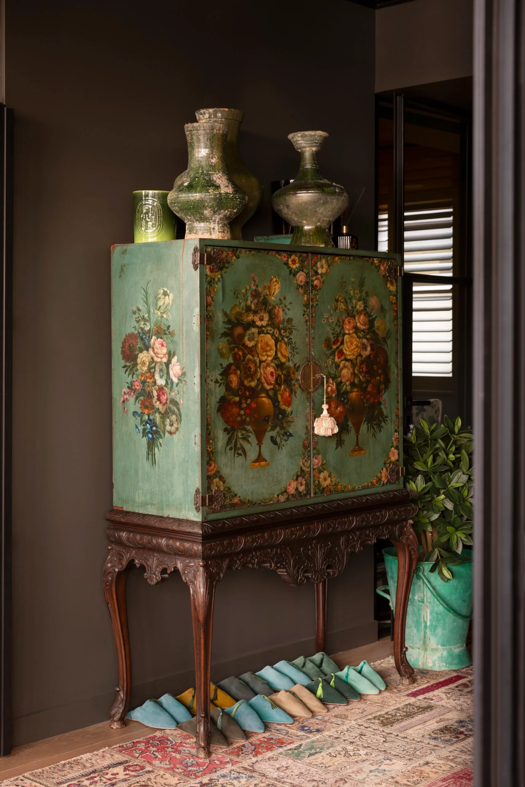 Antique cabinet with floral art, ornate vase decor on top, surrounded by potted plant and colorful slippers below.