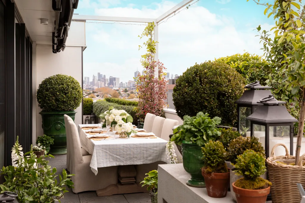 Outdoor terrace with a dining table set for six, surrounded by plants and city skyline in the background.