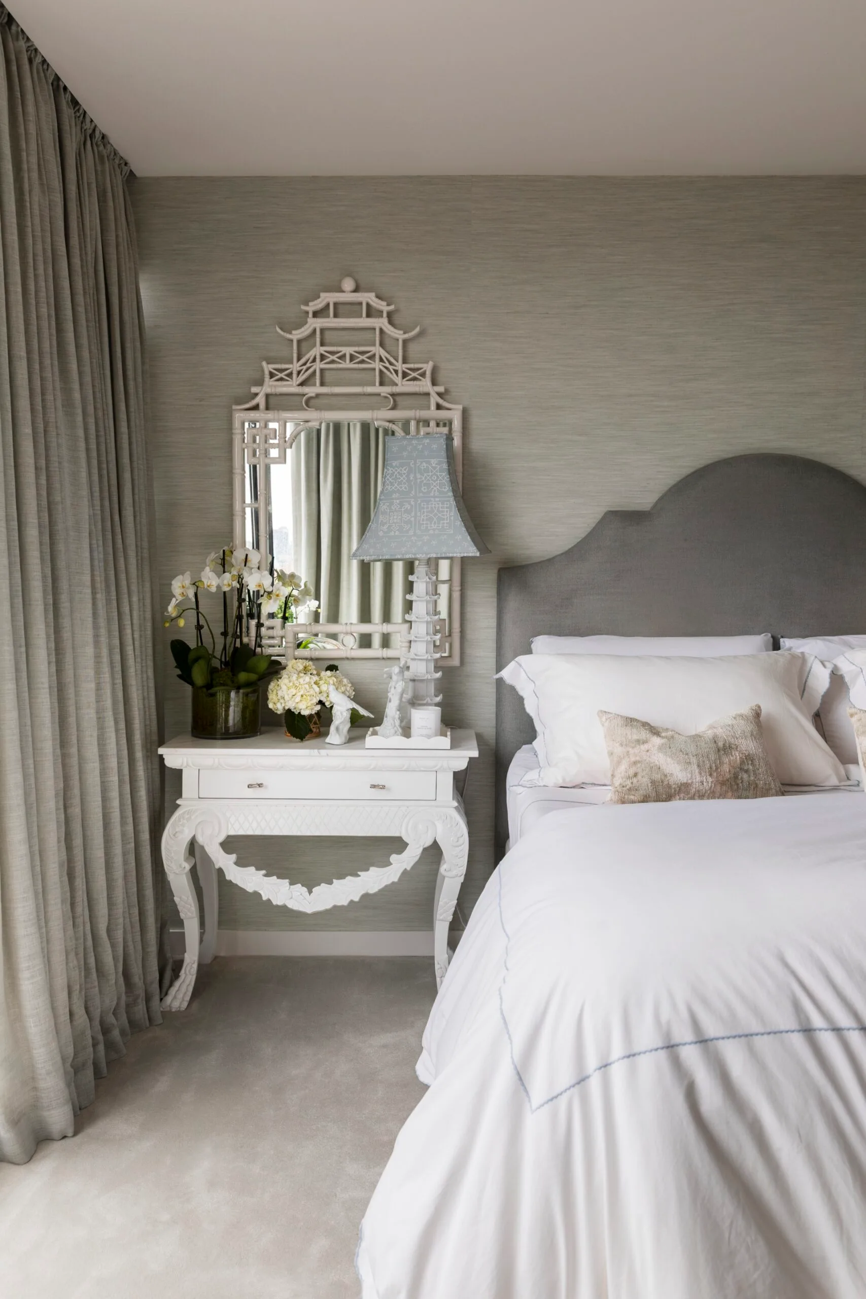Elegant bedroom with ornate white desk, decorative mirror, flowers, lamp, and a bed with gray headboard and white linens.