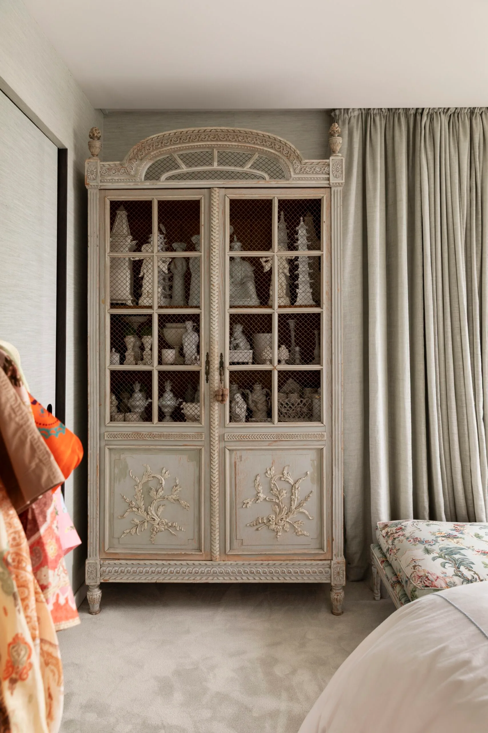 Antique ornate cabinet with decorative figurines inside, next to grey curtains and a floral cushion.