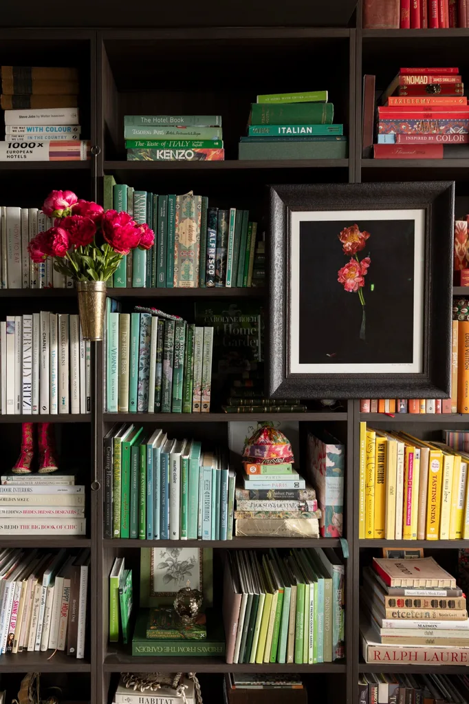 Bookshelf with color-coordinated books, a framed flower painting, red flowers in a vase, and decorative items.