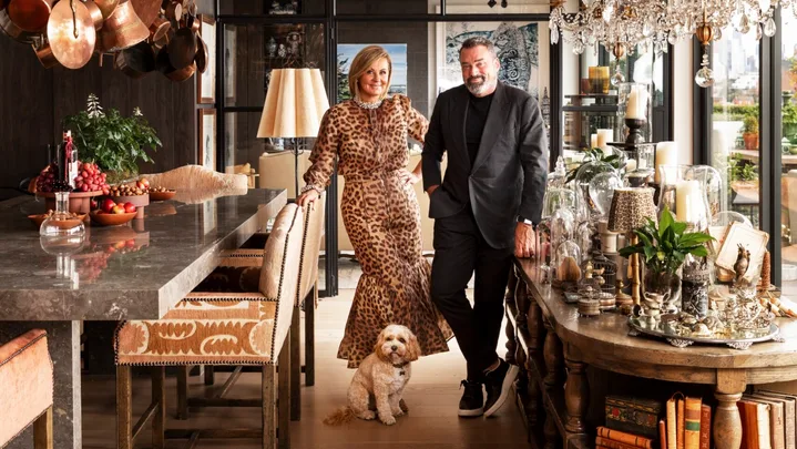 A stylish couple stands in a luxurious open plan kitchen and dining room with a small dog, surrounded by chandeliers and decor. The woman is Chyka Keebaugh, of Real Housewives of Melbourne fame and the man is her husband, Bruce Keebaugh.