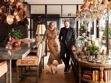 A stylish couple stands in a luxurious open plan kitchen and dining room with a small dog, surrounded by chandeliers and decor. The woman is Chyka Keebaugh, of Real Housewives of Melbourne fame and the man is her husband, Bruce Keebaugh.