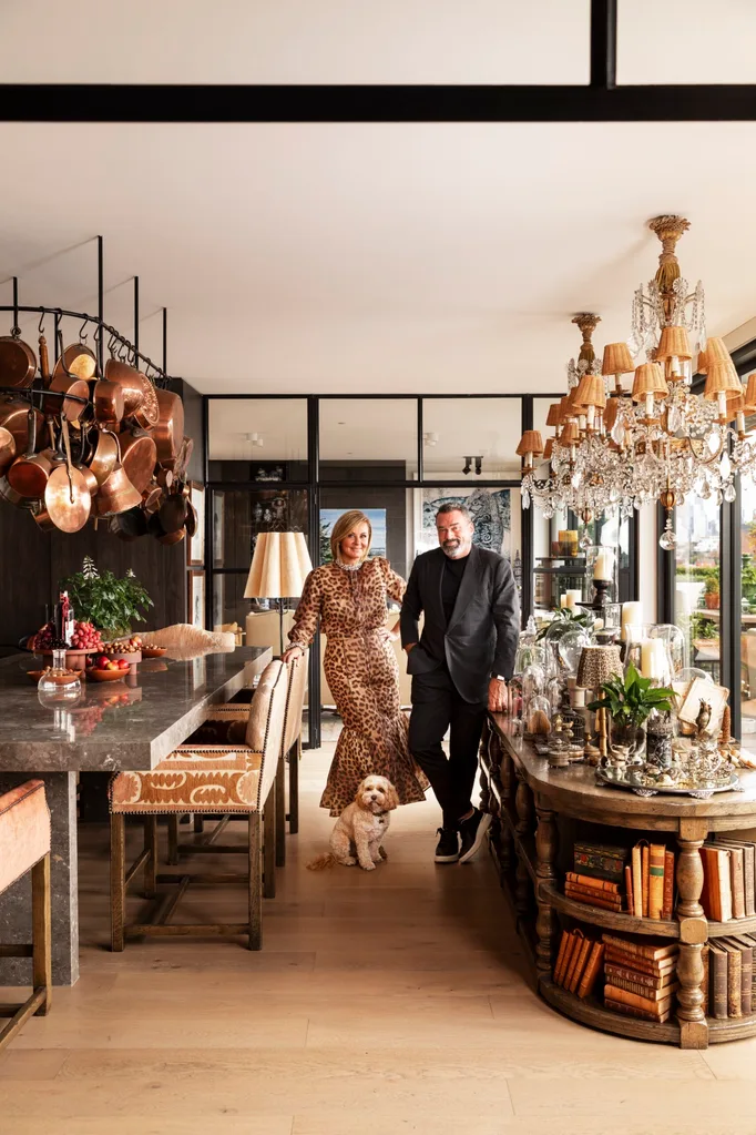 A stylish couple stands in a luxurious open plan kitchen and dining room with a small dog, surrounded by chandeliers and decor. The woman is Chyka Keebaugh, of Real Housewives of Melbourne fame and the man is her husband, Bruce Keebaugh.