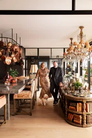 A stylish couple stands in a luxurious open plan kitchen and dining room with a small dog, surrounded by chandeliers and decor. The woman is Chyka Keebaugh, of Real Housewives of Melbourne fame and the man is her husband, Bruce Keebaugh.