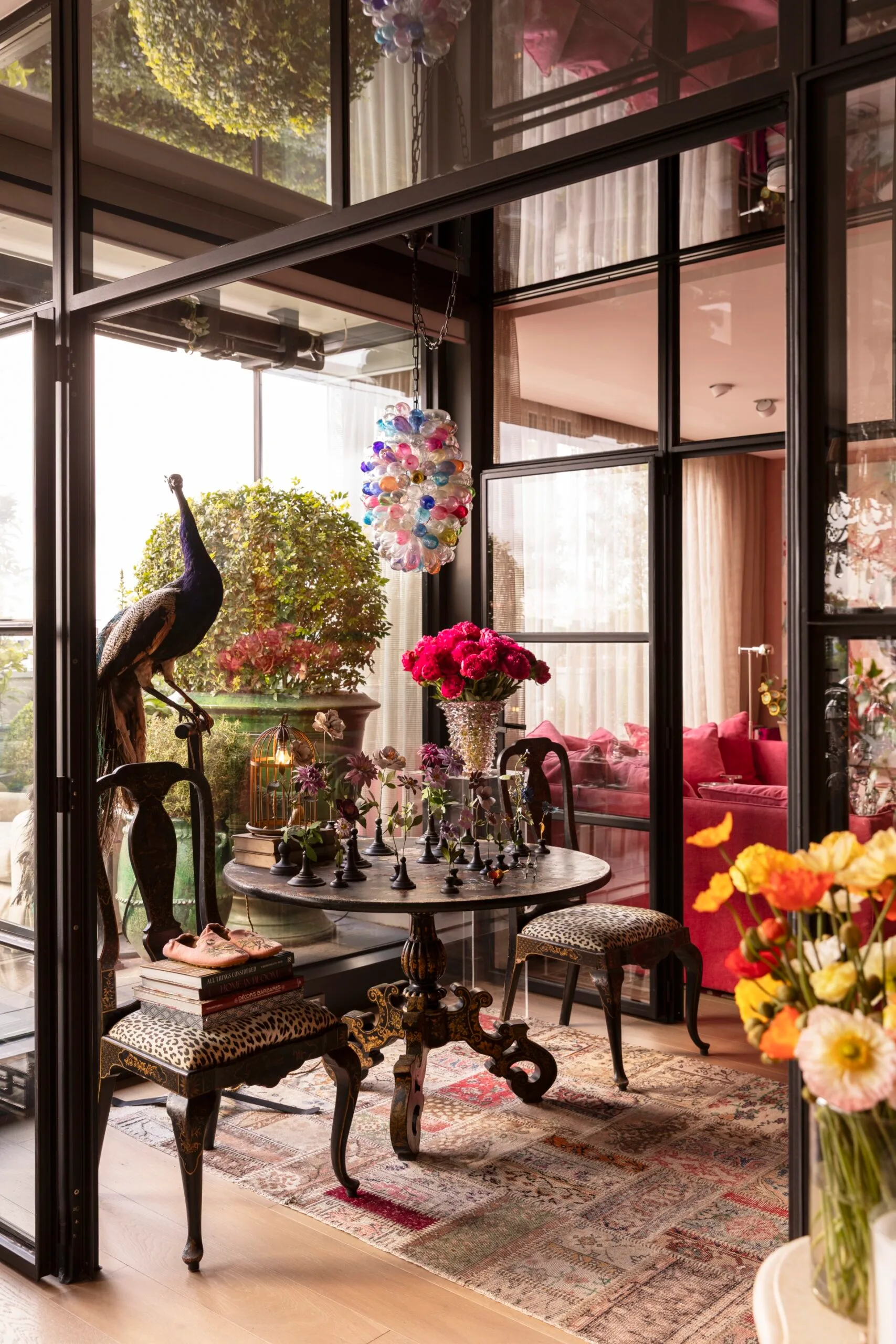 Elegant interior with a round table, chairs, colorful chandelier, peacock decor, and vibrant flowers by large windows.