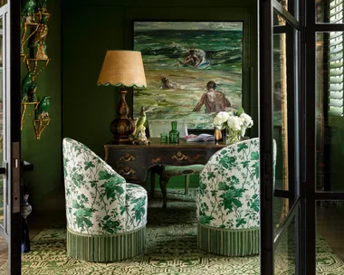 Elegant green room with floral chairs, painting, ornate desk, gold shelves with parrots, and a warm lamp. fringe decor trend