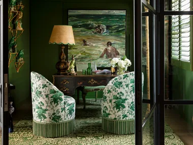 Elegant green room with floral chairs, painting, ornate desk, gold shelves with parrots, and a warm lamp. fringe decor trend
