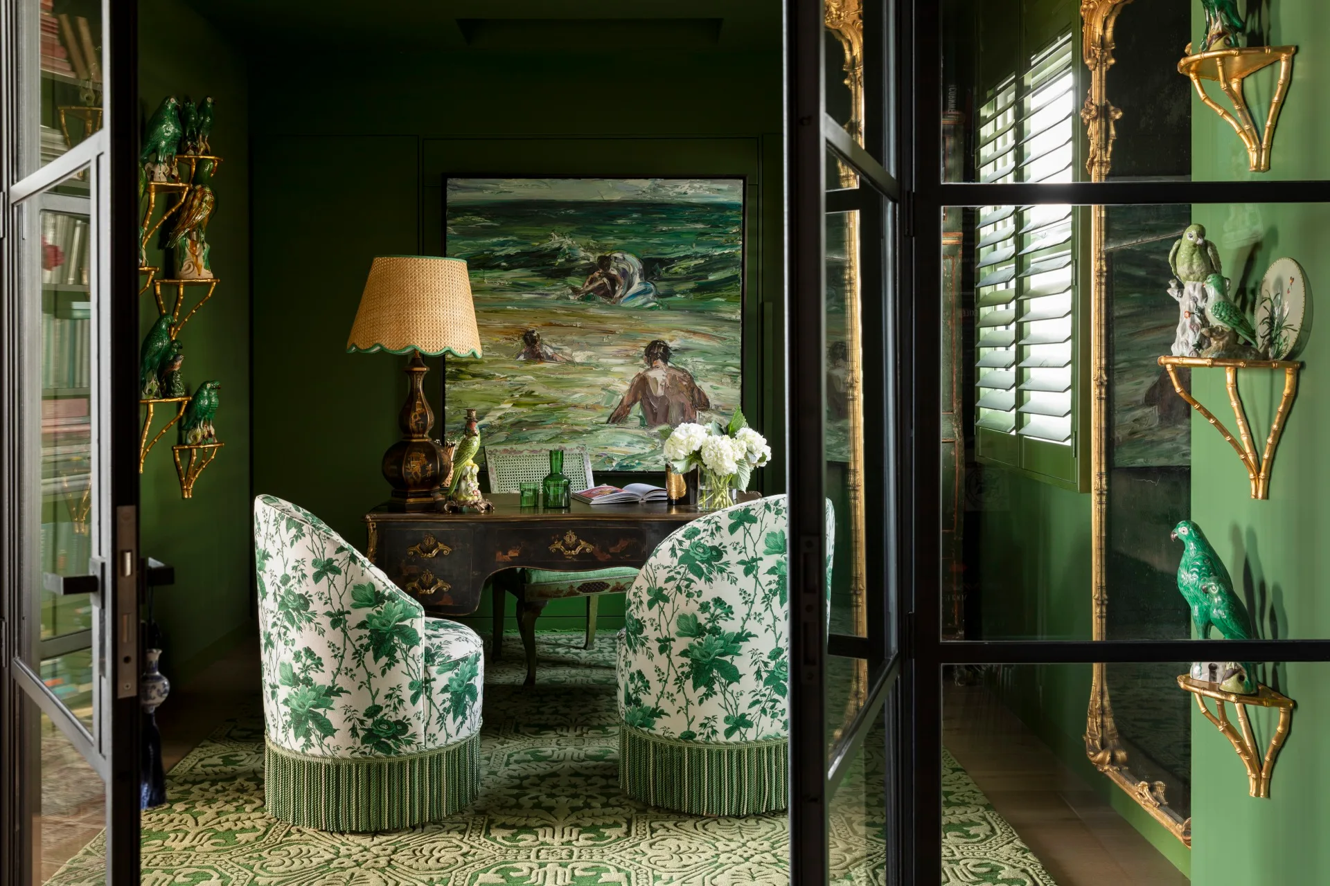 Elegant green room with floral chairs, painting, ornate desk, gold shelves with parrots, and a warm lamp. This is the library of TV star Chyka Keebaugh.
