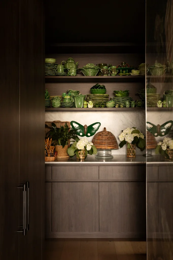 Shelves with green ceramic dishes, butterfly decor, potted plants, and flower arrangements in a dimly lit pantry. It is situated in the apartment penthouse home of Chyka Keebaugh from Real Housewives of Melbourne.